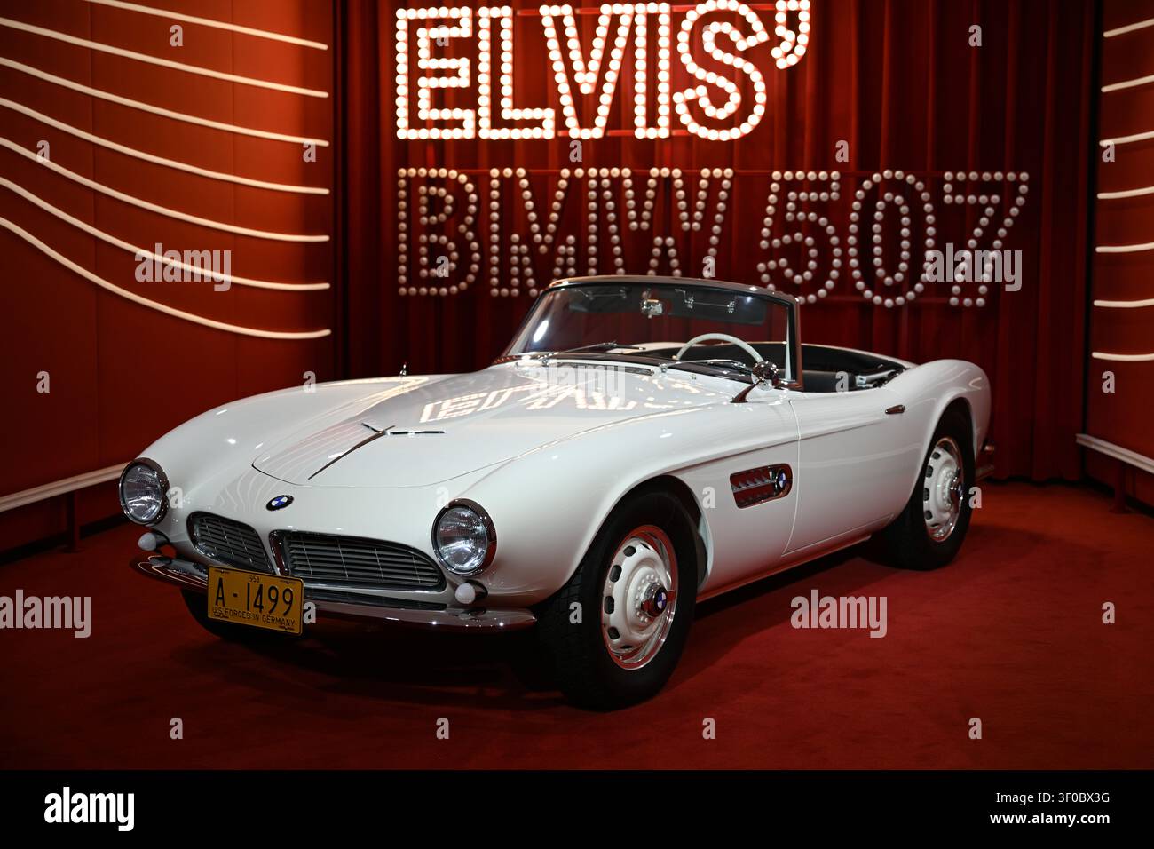 Città, Germania – 7 luglio 2026: Cabriolet BMW vintage esposto in una mostra con cartello luminoso 'ELVIS' e sfondo di tende rosse. Foto Stock