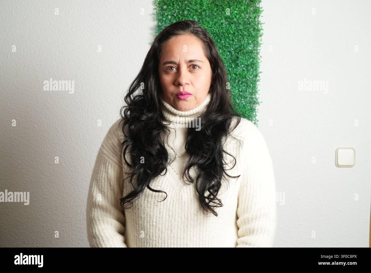 Donna in piedi davanti a una siepe artificiale verde. Indossa un maglione a collo alto color crema. I capelli sono lunghi, scuri e mossi. Il muro dietro di lei è semplice Foto Stock
