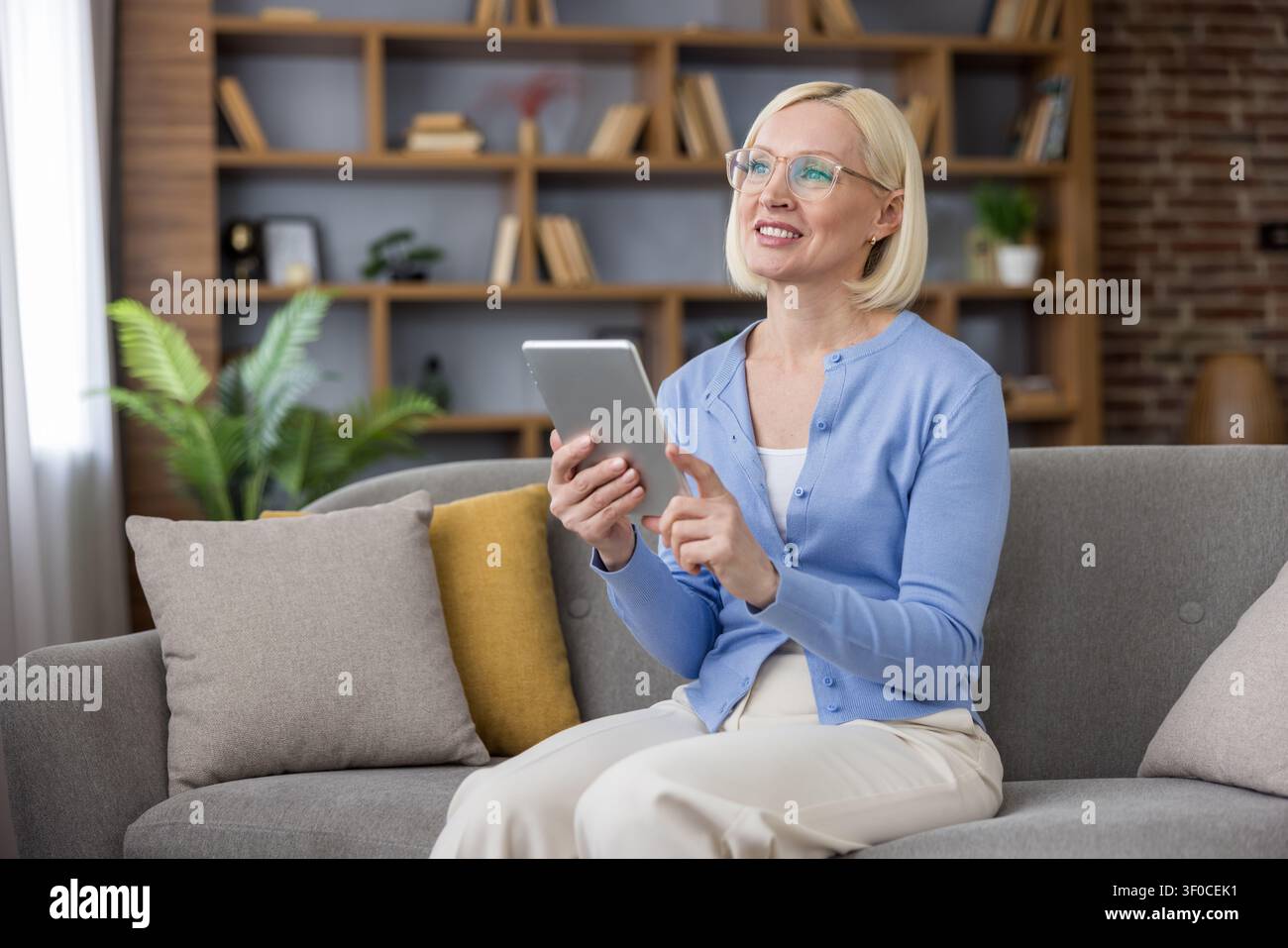 Donna anziana felice seduta su un comodo divano, sorride mentre guarda verso l'alto e tiene un tablet digitale, connettendosi con la tecnologia e l'istruzione online a casa Foto Stock