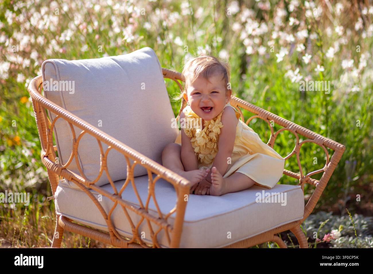 Ritratto di una bambina seduto su una sedia nel giardino estivo. Un bambino allegro e divertente si rilassa all'aperto tra fiori in fiore Foto Stock
