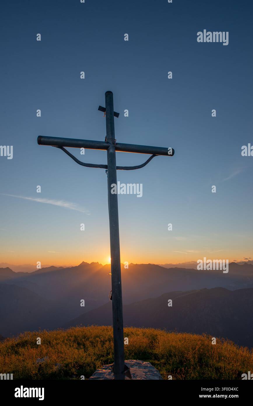 I raggi del sole al tramonto filtrano da dietro la croce creando un effetto di luce divina e spiritualità che avvolge le vette circostanti Foto Stock