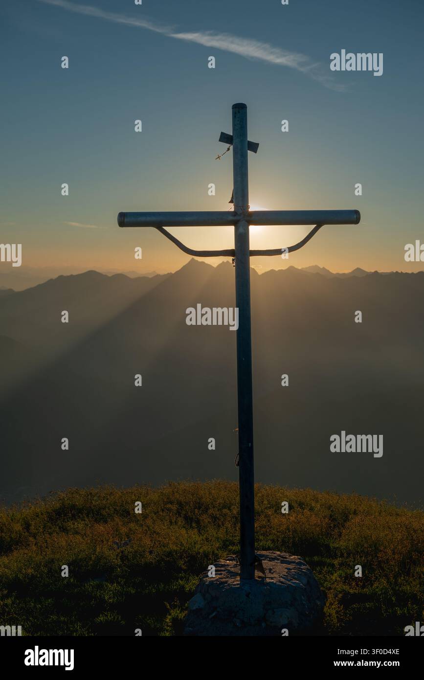 I raggi del sole al tramonto filtrano da dietro la croce creando un effetto di luce divina e spiritualità che avvolge le vette circostanti Foto Stock
