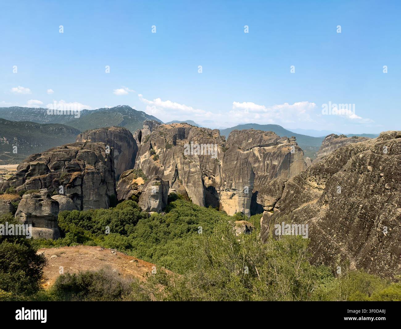 Kalambaka, Grecia – 24 luglio 2024: Paesaggio delle Meteore. Gruppo di torri rocciose nere e gialle in piedi, inserite nella natura verdeggiante di alberi e cespugli sotto Foto Stock