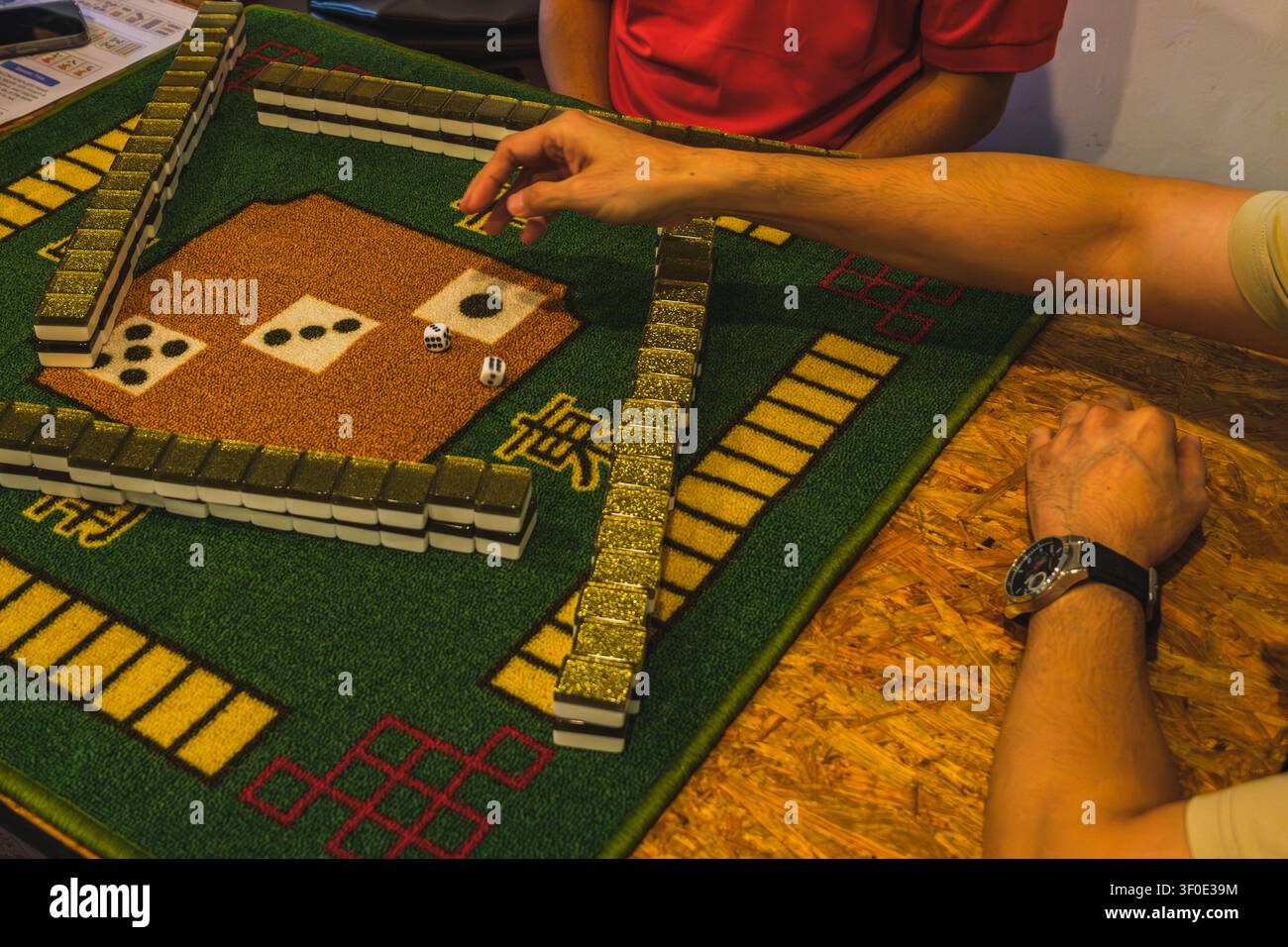 Un primo piano ad angolazione alta di giocatori che preparano una partita di Mahjong, con muri di piastrelle impilati su un tappeto verde e una coppia di dadi lanciati sul tavolo Foto Stock