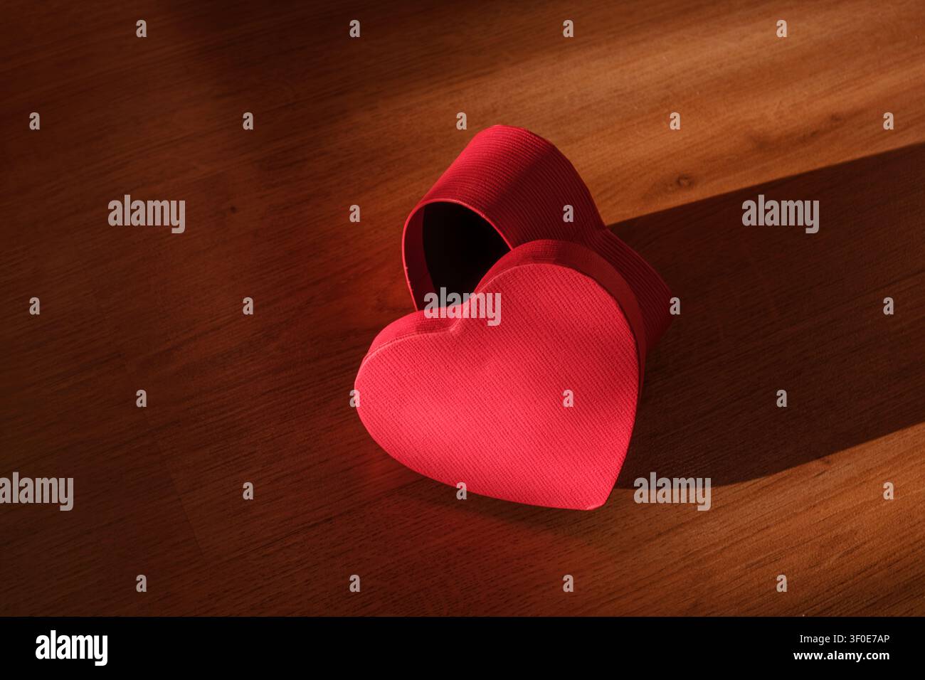 Una scatola rossa a forma di cuore con coperchio aperto posizionata su una superficie di legno alla luce calda del sole. Simboleggia amore, romanticismo e affetto. Per temi come San Valentino Foto Stock