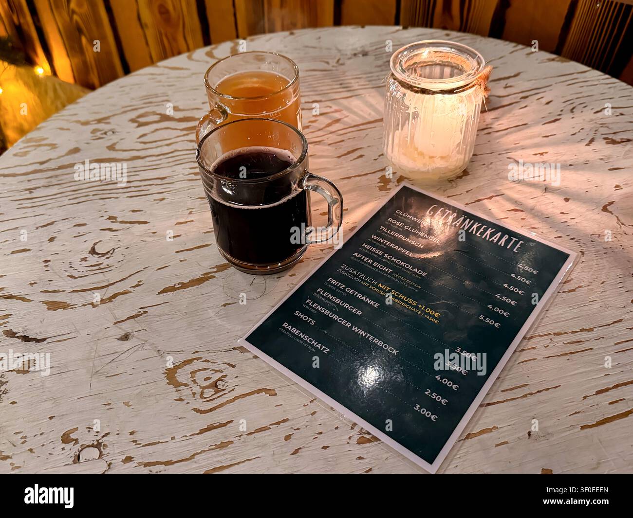 Tre bicchieri di punch al vino caldo su un tavolo di legno rustico con un menù che mostra opzioni di bevande e prezzi illuminati da una candela in un ambiente accogliente Foto Stock