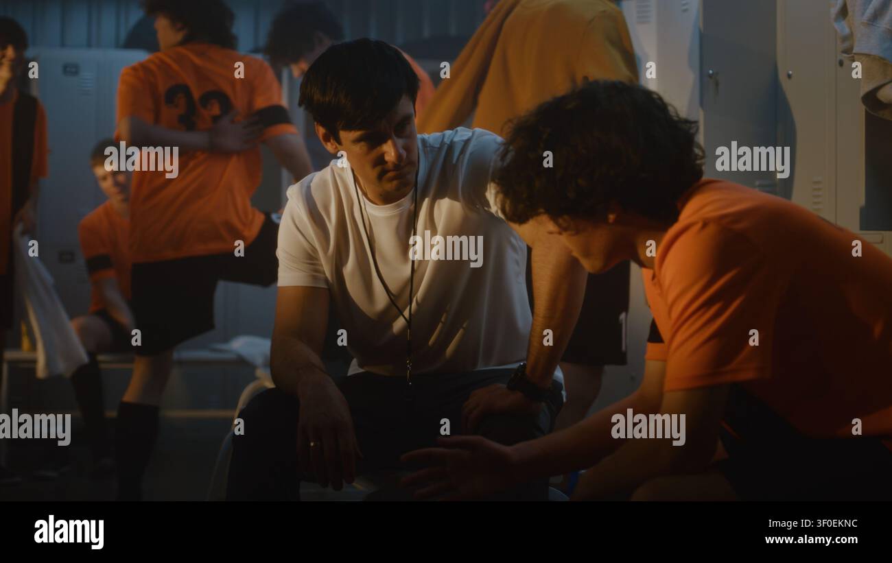L'allenatore di calcio si prende un momento per motivare uno dei giocatori adolescenti nello spogliatoio mentre i compagni di squadra cambiano i vestiti e si riprendono nelle vicinanze. Mentorship, leadership, relazioni forti che modellano squadre di successo. Foto Stock