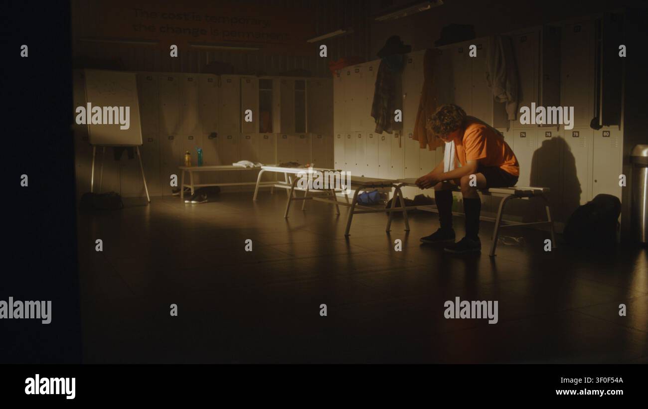 Allenatore di calcio si avvicina a un adolescente deluso nello spogliatoio vuoto dopo una partita difficile, offrendo incoraggiamento e sostegno emotivo. Resilienza, mentorato, lato umano dello sport competitivo. Foto Stock