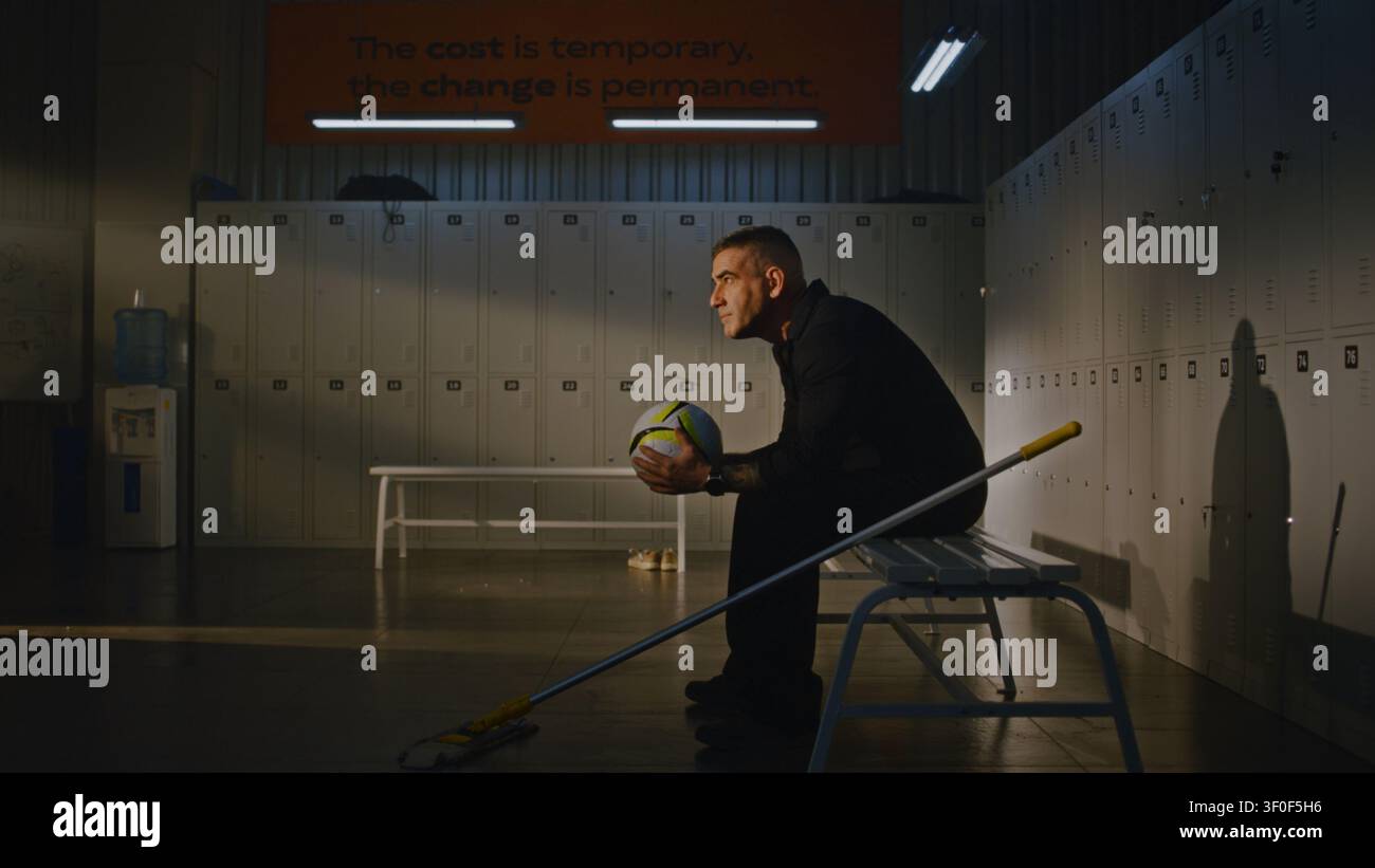 Addetto stanco fa una pausa dallo spazzare lo spogliatoio sportivo. L'uomo si siede su una panchina, raccoglie un pallone da calcio lasciato dalla squadra e lo tiene con entrambe le mani, riflettendo sulla gioventù con un senso di nostalgia. Foto Stock