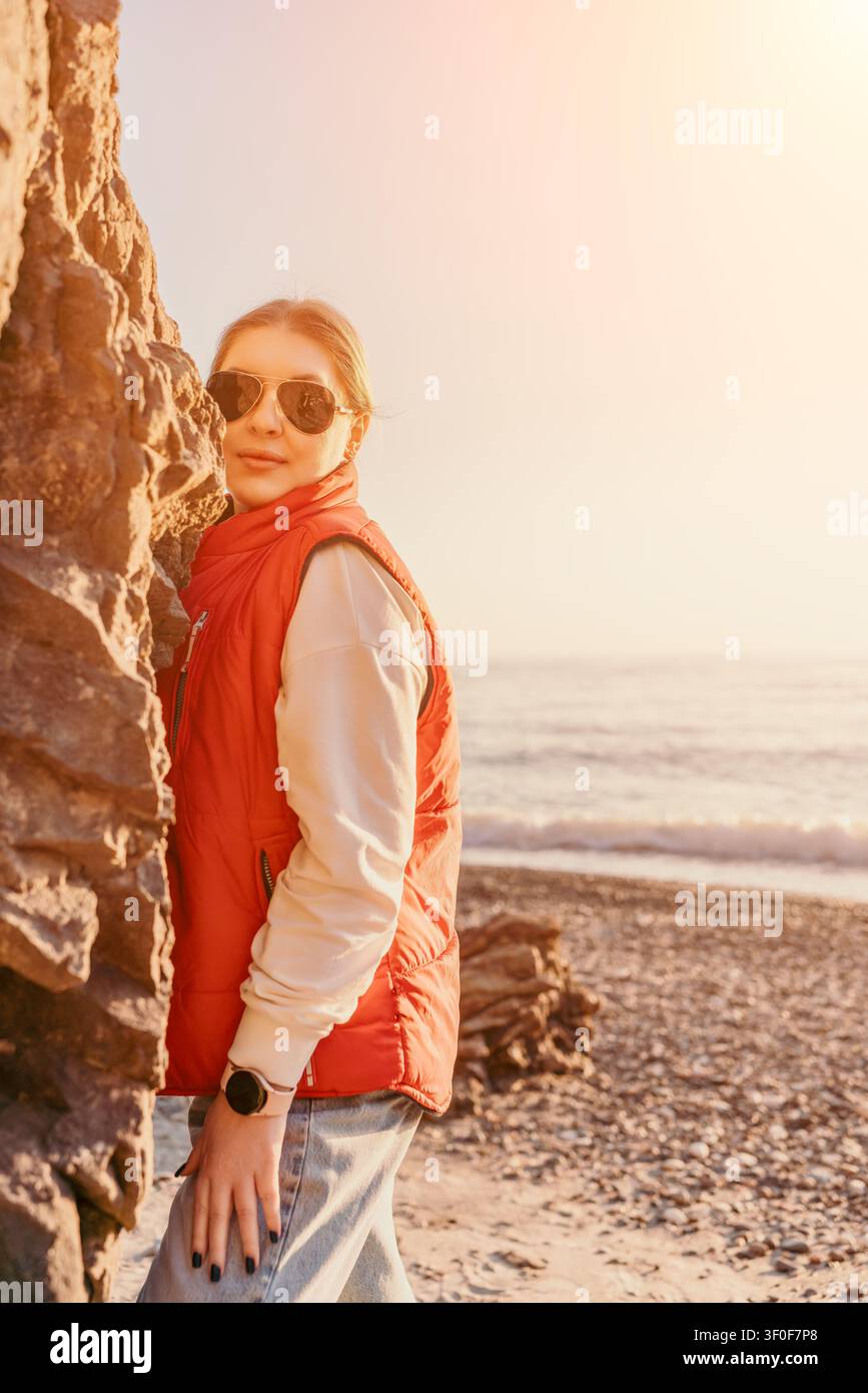 Donna viaggiatrice sorridente in un giubbotto rosso in piedi su una roccia su una spiaggia di ciottoli al tramonto con spazio per il testo Foto Stock