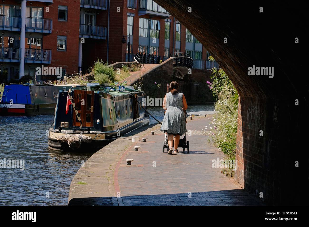 Una donna con un passeggino che cammina lungo il sentiero del canale oltre le chiatte ormeggiate a Birmingham, UK Foto Stock