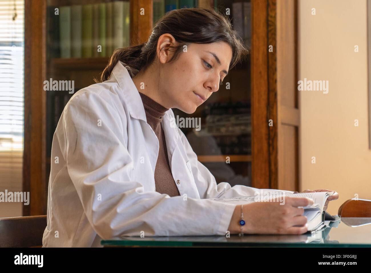 Giovane scienziata in camice bianco che legge con attenzione da un grande libro aperto a una scrivania di vetro, con una libreria antica in legno piena di libri nell' Foto Stock