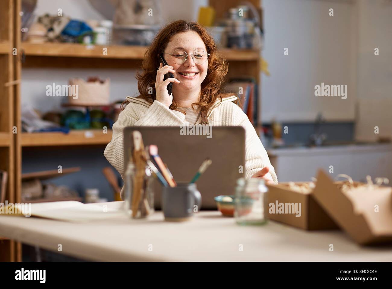 Proprietaria di uno studio di ceramica che sorride al telefono mentre lavora su un laptop e imballa gli ordini Foto Stock