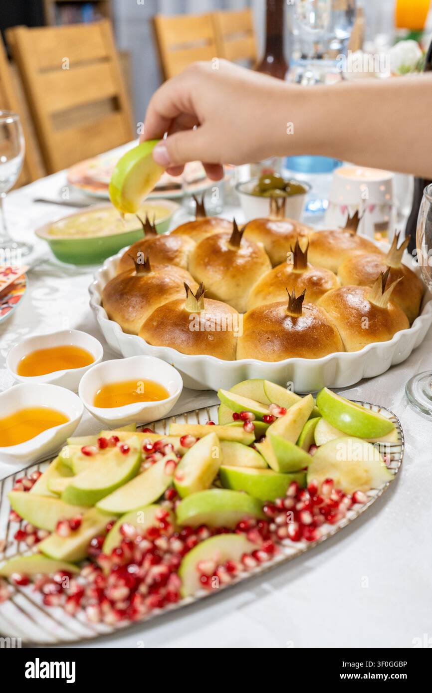 Le persone si radunano intorno a un tavolo per Rosh Hashanah. Una mano immerge una fetta di mela nel miele. Frutta fresca e pane dolce fanno parte del pasto, mostrando usanze importanti. Foto Stock