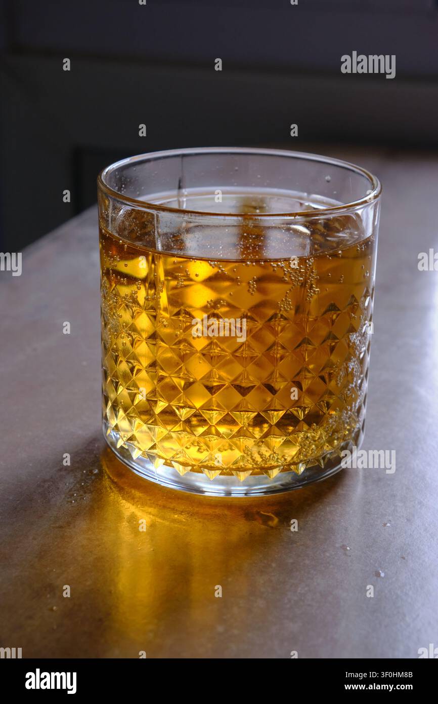 Bicchiere di whisky dorato con cubetti di ghiaccio appoggiato su un tavolo di legno scuro. Il ricco liquido ambrato riflette una luce ambientale calda, creando riflessi morbidi Foto Stock