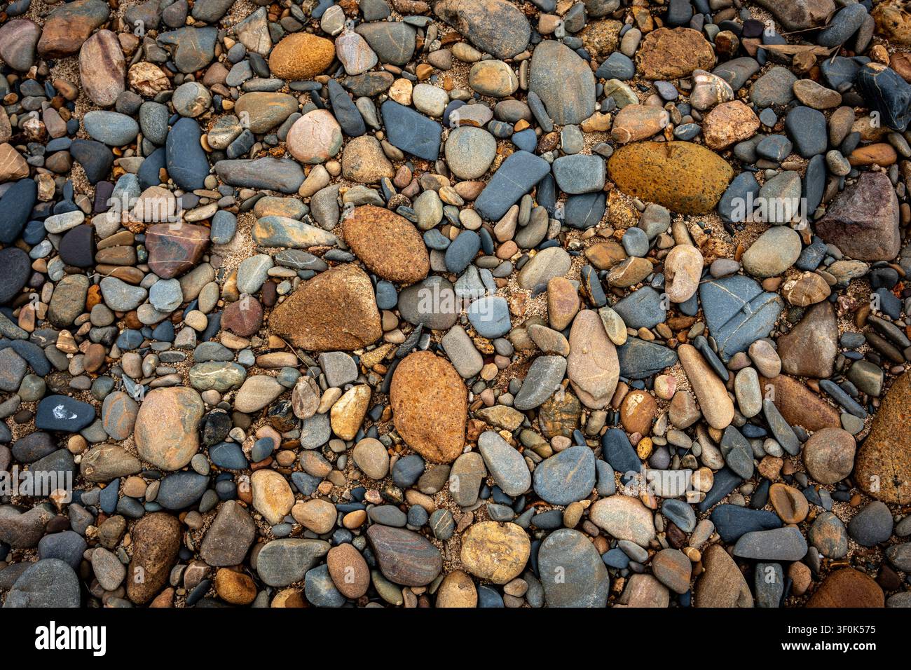 Una visione a tutto campo, dall'alto, di ciottoli di fiume lisci e colorati e sabbia. Uno sfondo con texture naturale e terrosa con varie sfumature di marrone, grigio, blu Foto Stock