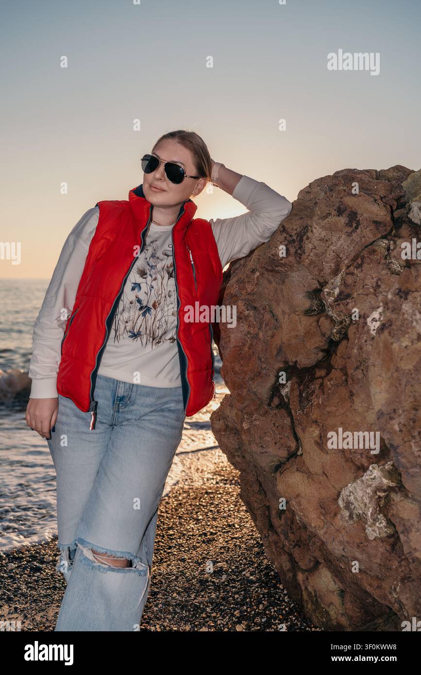 Donna in posa su una spiaggia al tramonto contro un grande scoglio costiero durante l'ora dorata, indossando un piumino rosso e jeans Foto Stock