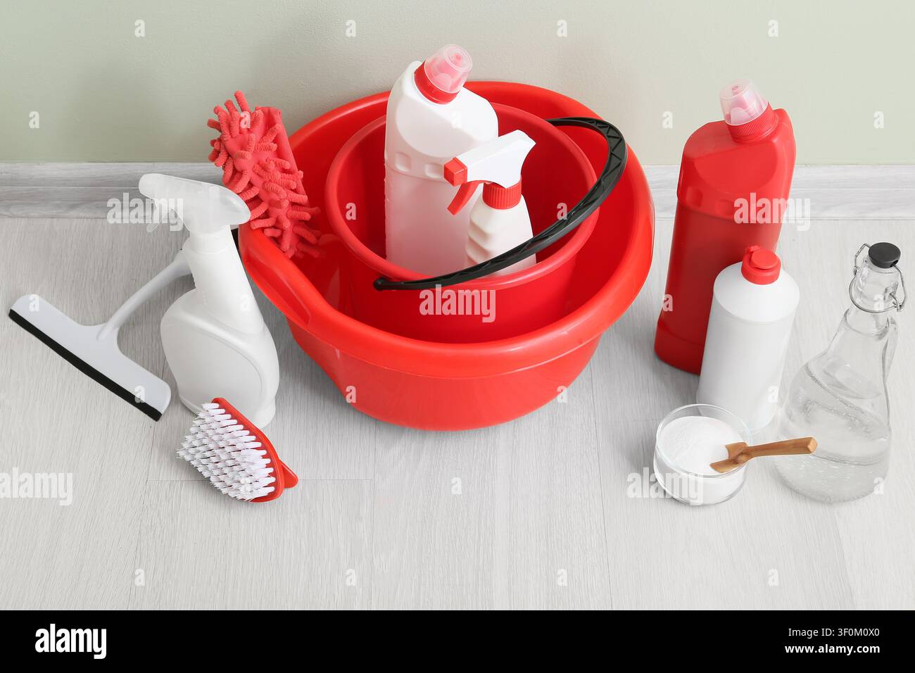 Prodotti e strumenti per la pulizia tra cui secchi rossi, flaconi spray e spazzole su un pavimento di legno chiaro Foto Stock