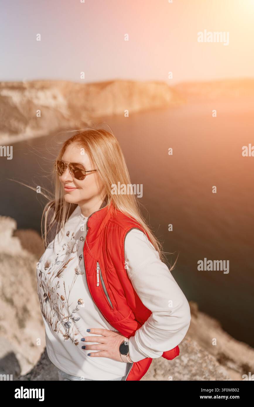 Donna viaggio costiero donna bionda che indossa un giubbotto rosso e occhiali da sole in piedi su una scogliera panoramica che si affaccia sul mare al tramonto Foto Stock