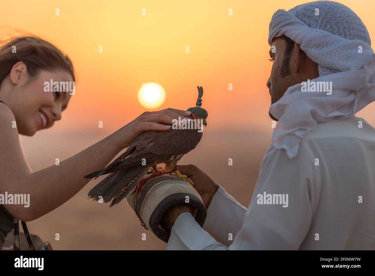 Un falconiere emirati vestito in modo tradizionale tiene un falco incappucciato su un guanto di pelle mentre una turista accarezza delicatamente l'uccello rapace contro il. Foto Stock