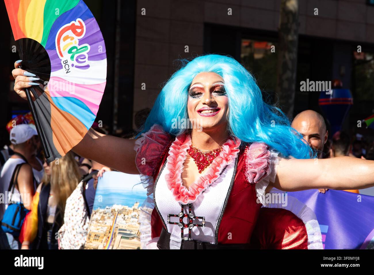 Una drag queen con una parrucca blu brillante e un abito a balze rosa danza allegramente al Barcelona Pride, sventolando un ventaglio arcobaleno "Pride BCN". Foto Stock