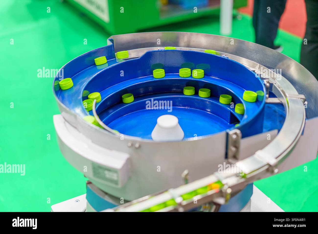 Un primo piano di un alimentatore vibrante industriale che smista tappi di bottiglie in plastica verde su una linea di produzione, mostrando la produzione automatizzata e il packa Foto Stock