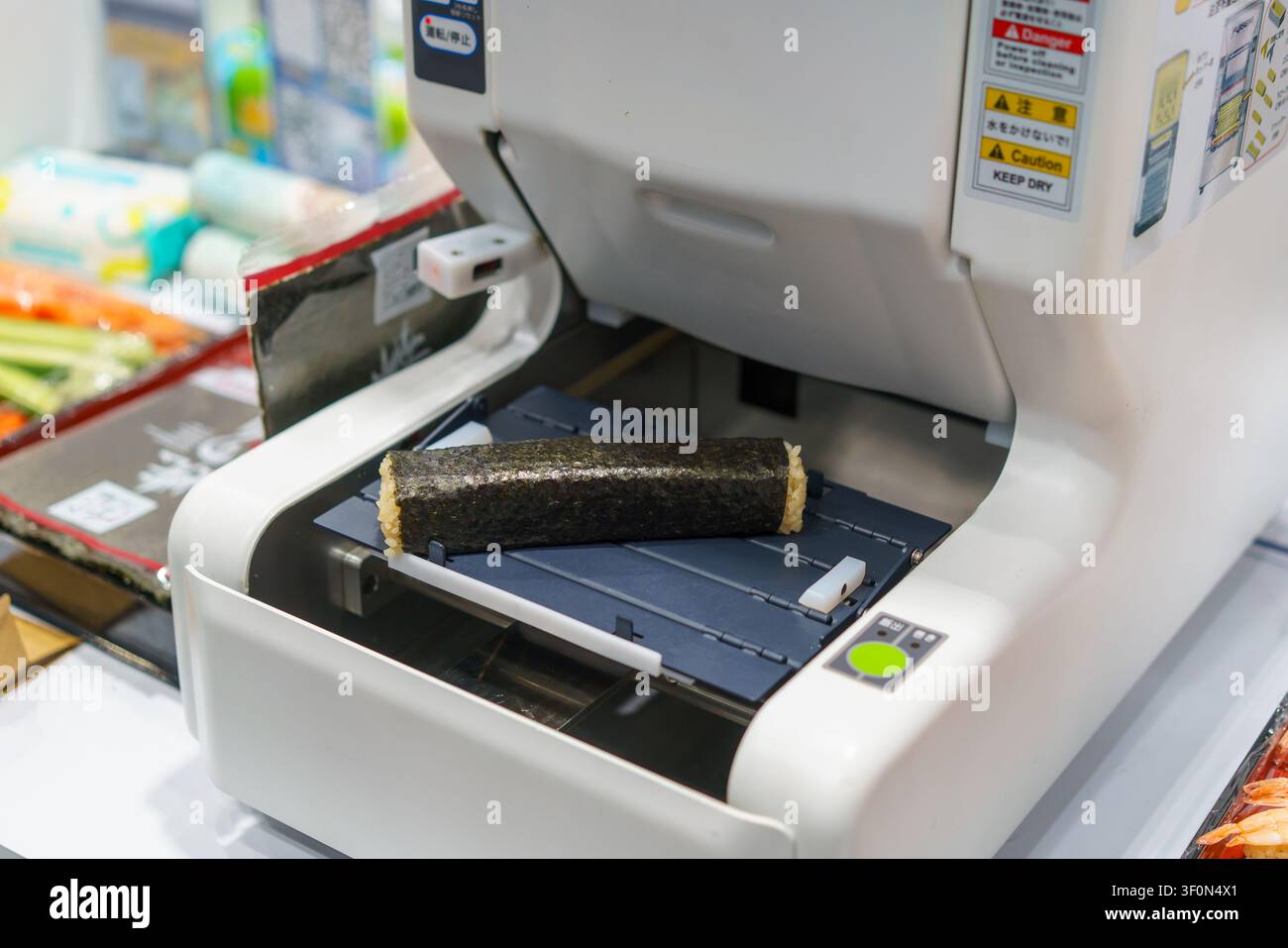 Primo piano di una macchina commerciale automatizzata per la preparazione del sushi che distribuisce un maki appena arrotolato avvolto in alghe nori sul suo vassoio. Ideale per ristoranti moderni Foto Stock