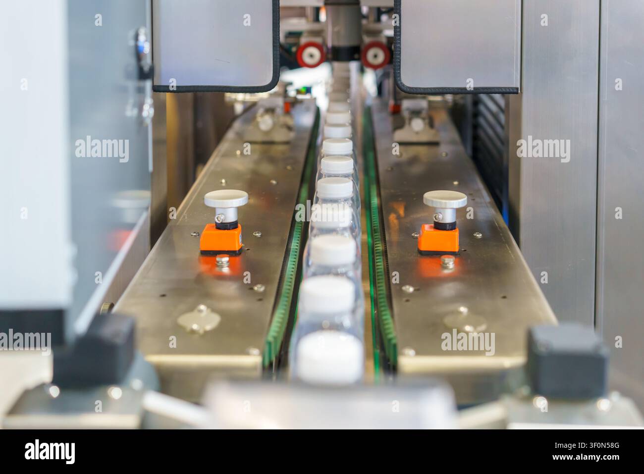 Bottiglie di plastica trasparenti vuote con tappi bianchi che avanzano su un nastro trasportatore in acciaio inossidabile all'interno di uno stabilimento di produzione industriale automatizzato. Foto Stock