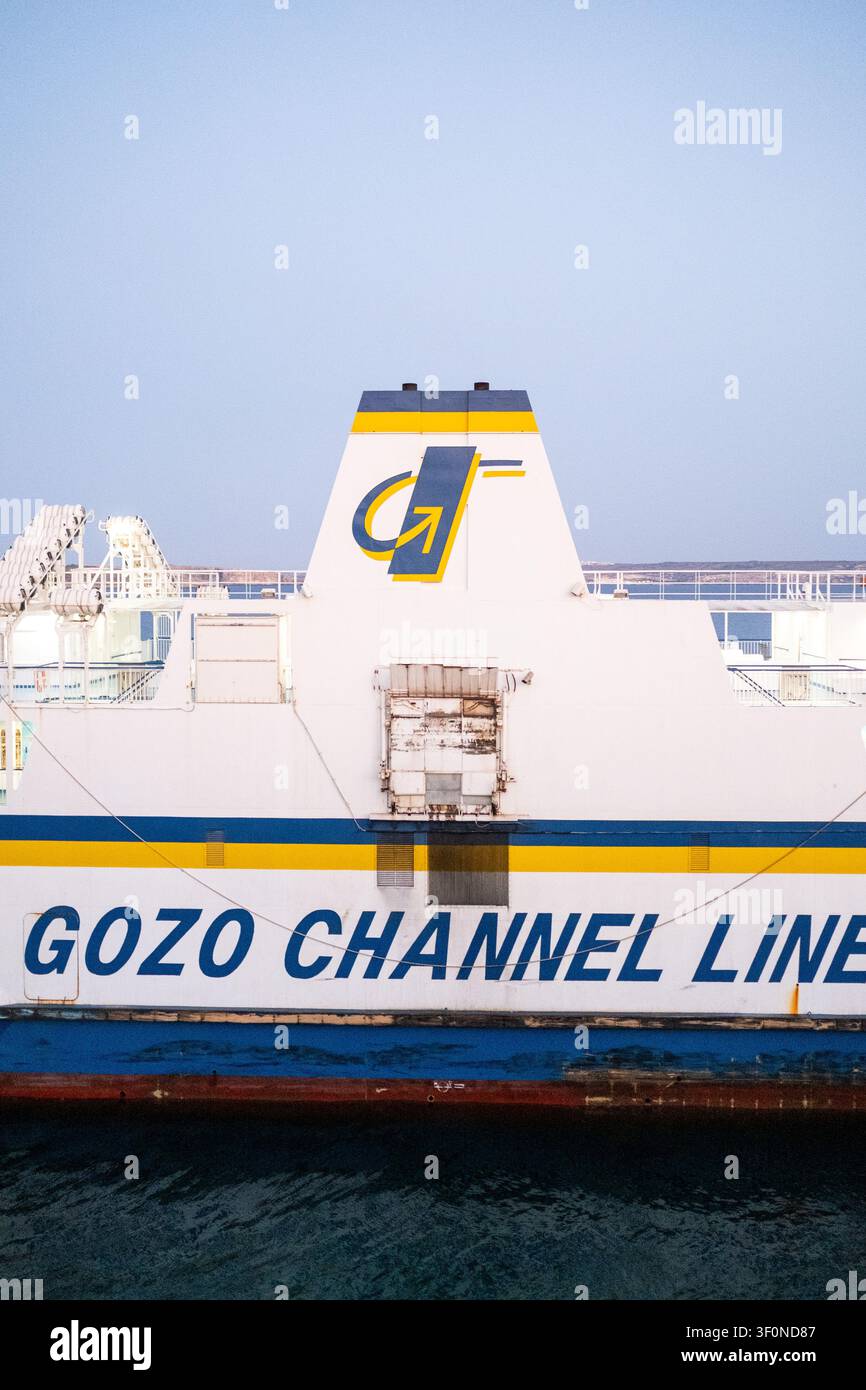 Vista ravvicinata del lato di una nave della Gozo Channel Line al porto di Mgarr, con il nome dell'azienda, i segni blu e gialli, i ponti passeggeri illuminati, li Foto Stock