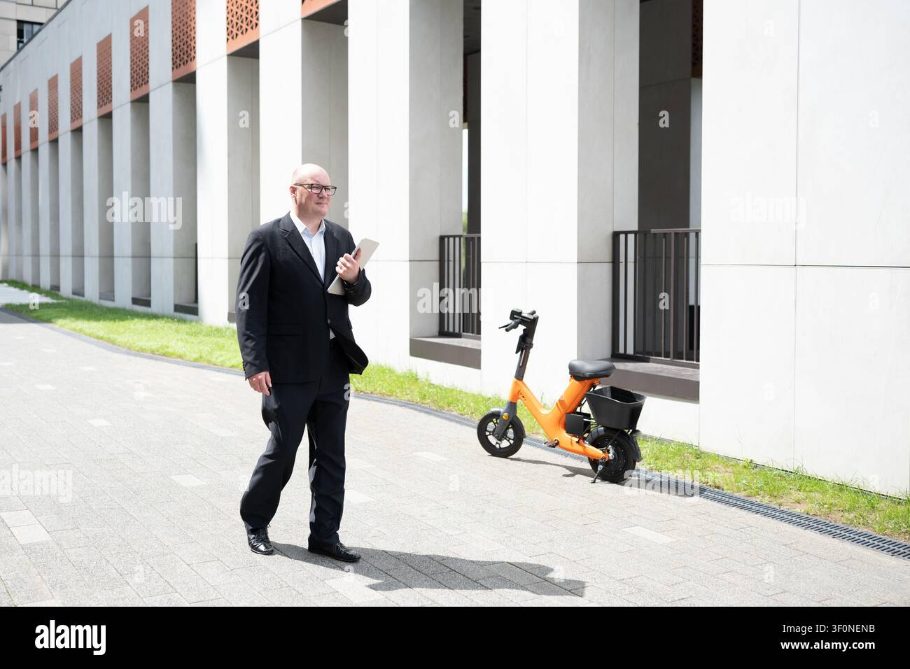 Uomo d'affari caucasico che cammina con un laptop vicino a una bici elettrica fuori da un edificio di uffici moderno. Mobilità aziendale, pendolarismo, trasporto urbano e città intelligente Foto Stock