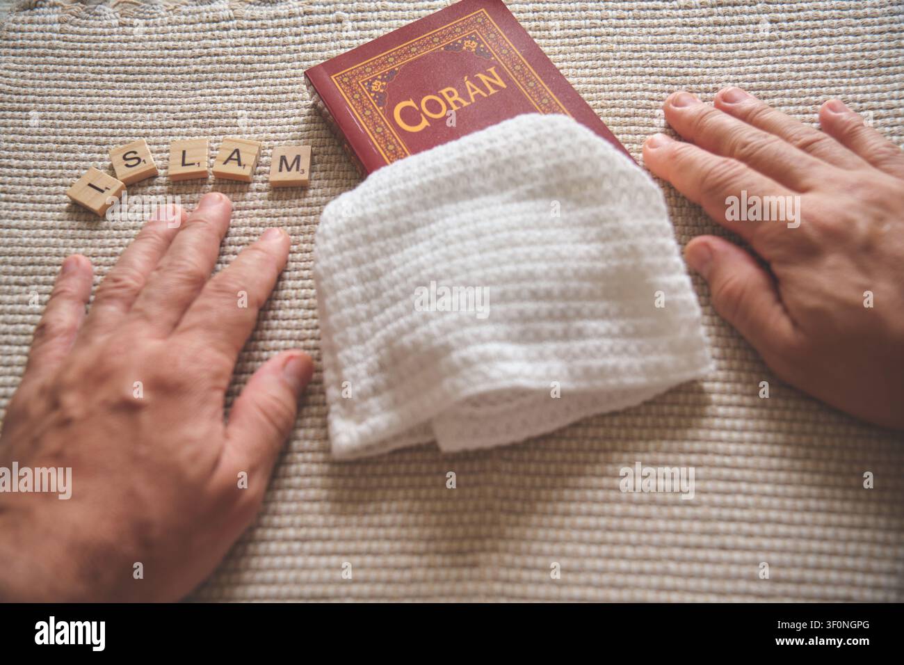 Mani appoggiate accanto a un libro del Corano, un cappello da preghiera kufi e la parola "Islam" scritta con tessere di Scrabble Foto Stock