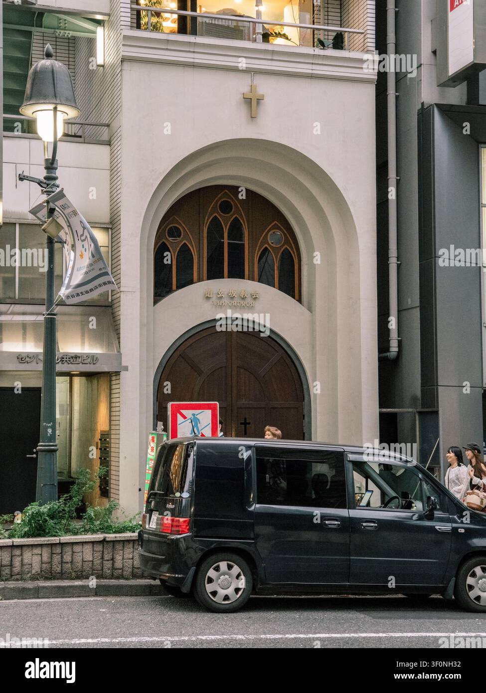 Facciata stretta di cappella cristiana con croce e porta in legno ad arco tra gli edifici di Dogenzaka, Shibuya, Tokyo, con un minivan nero parcheggiato Foto Stock