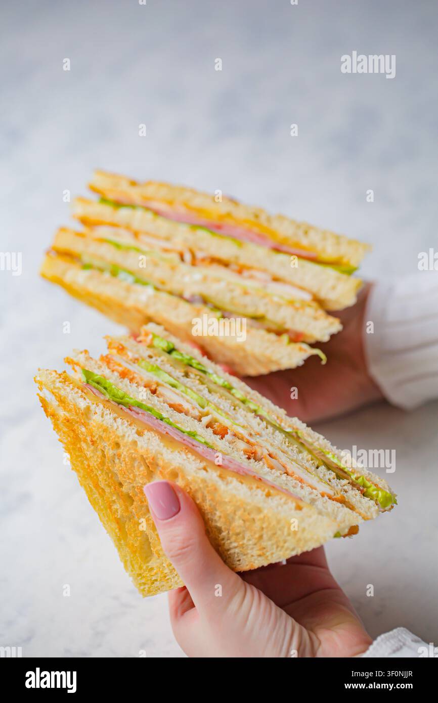 Due mani tengono due panini impilati ripieni di strati di carne, lattuga e formaggio. La cucina ha un design semplice con uno sfondo chiaro. Foto Stock