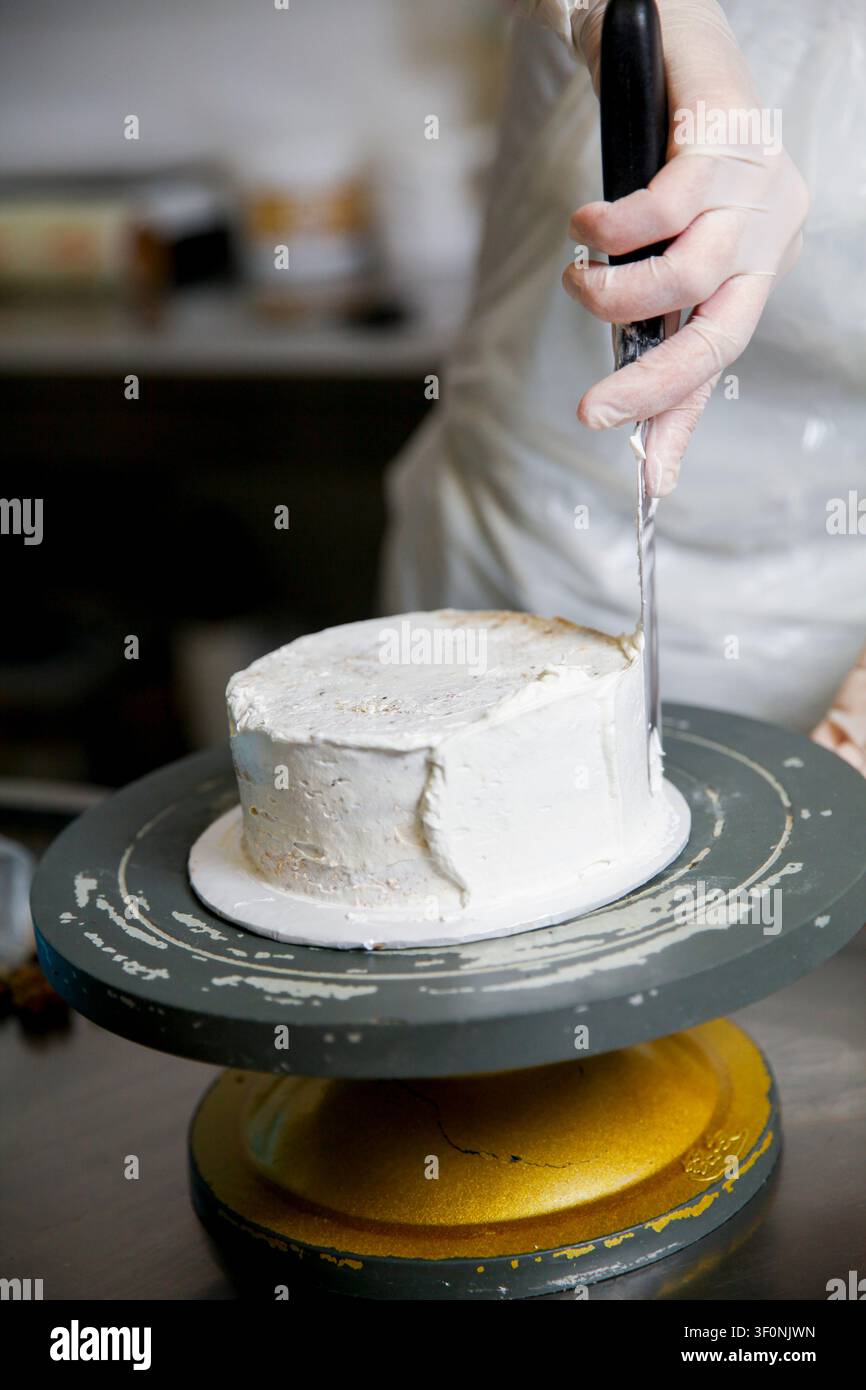 Un fornaio usa un coltello per levigare la glassa su una torta mentre gira su un piano girevole. La cucina è indaffarata con ingredienti sparsi intorno. Foto Stock