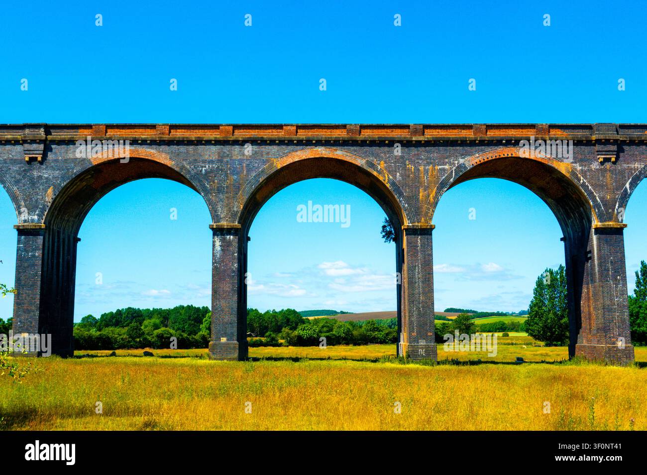 Viadotto ferroviario in mattoni vittoriano con tre archi che incorniciano la campagna inglese verde e l'erba dorata delle praterie estive, Inghilterra, Regno Unito. Foto Stock