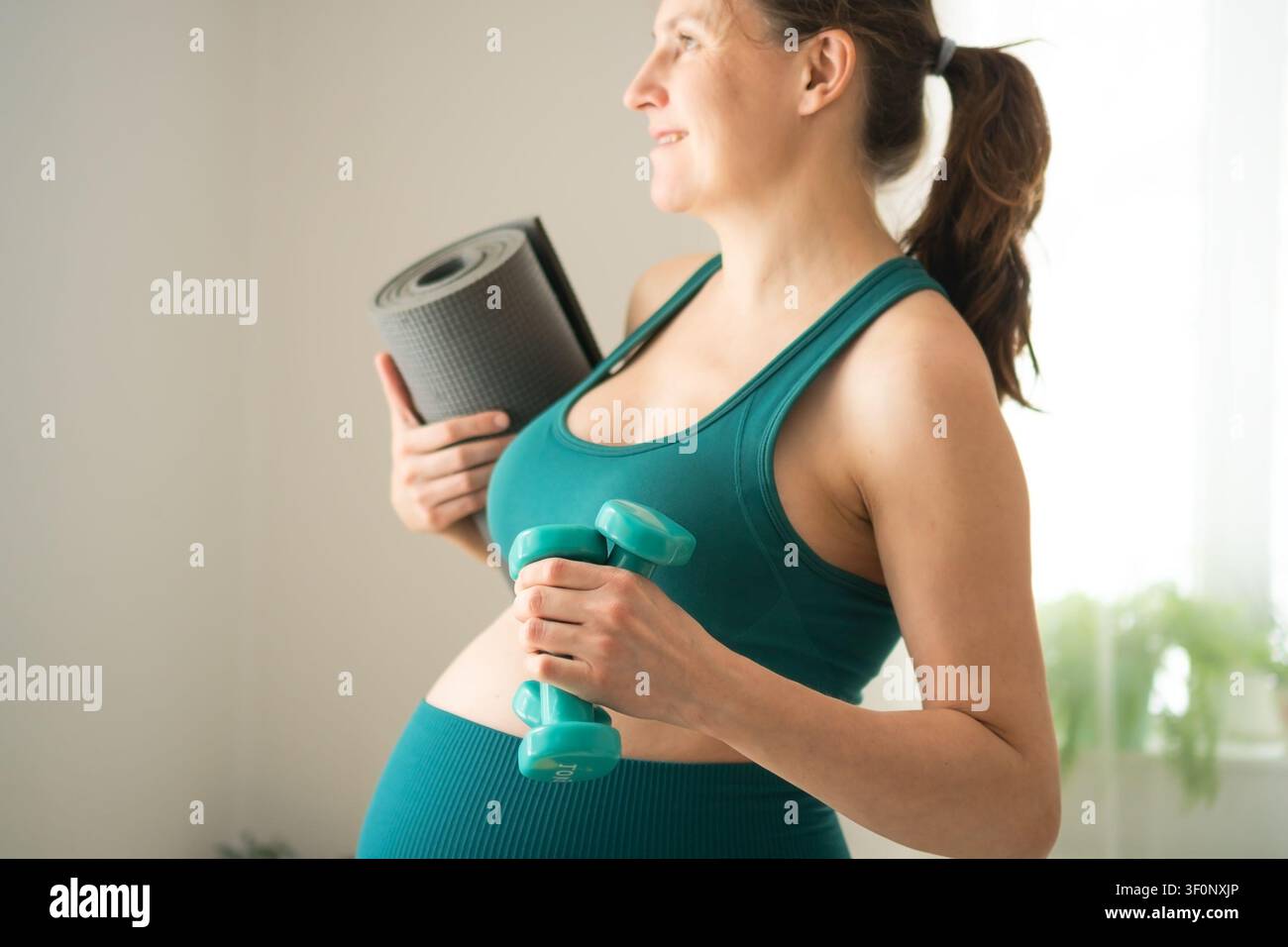 Donna incinta in comoda activewear, vista di lato per l'allenamento prenatale. La futura madre indossa un moderno abbigliamento fitness da maternità con manubrio. La futura mamma rimane sicura di sé e alla moda durante l'esercizio Foto Stock