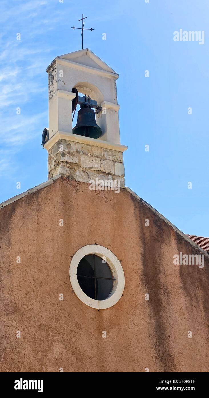 Primo piano di un campanile rustico con una campana in metallo e una croce in cima, caratterizzato da una finestra rotonda e da un esterno ruvido sotto un cielo blu terso. Foto Stock