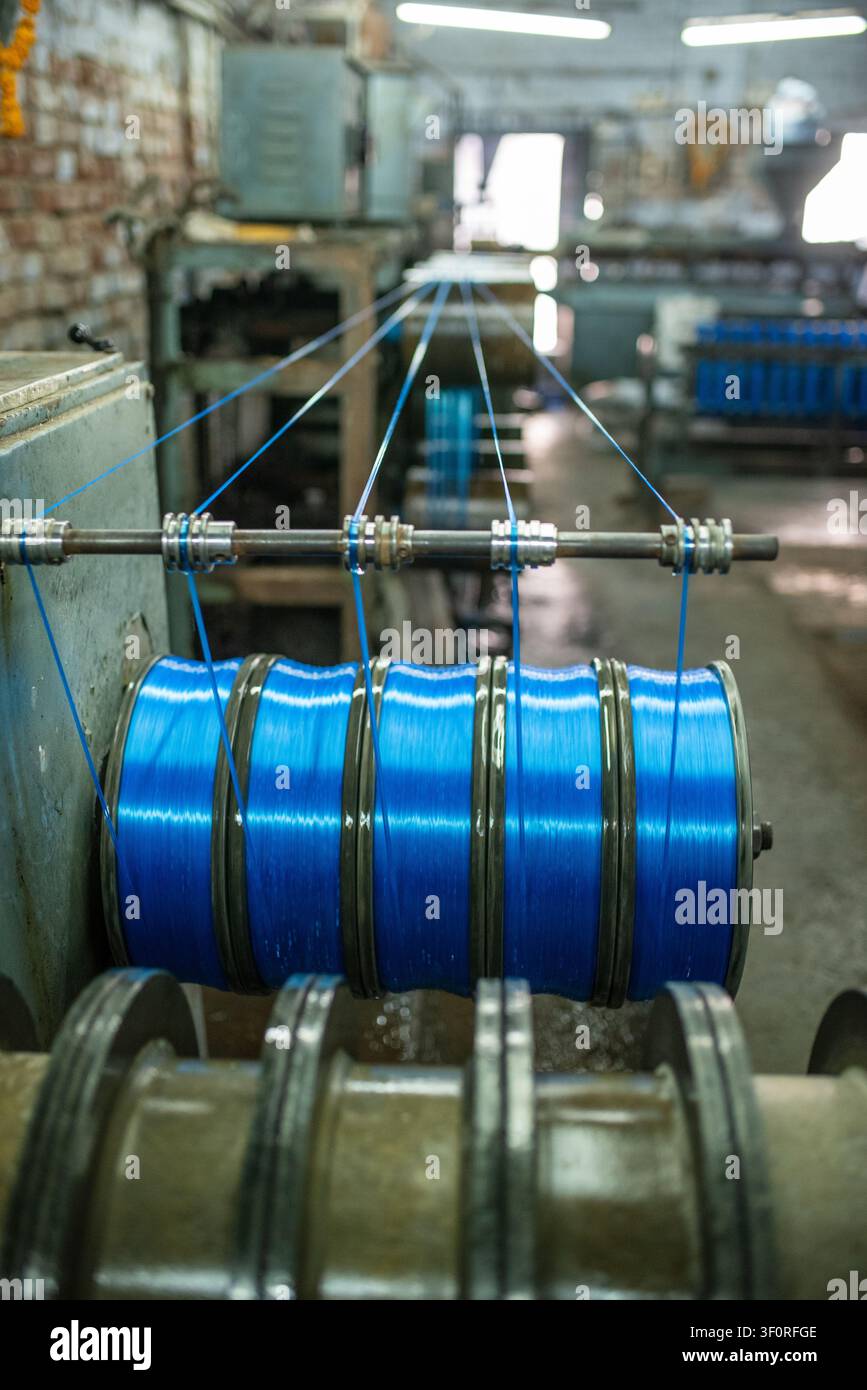 Vista ravvicinata di grandi bobine metalliche che avvolgono un filamento di plastica blu lucido in una fabbrica di produzione a Bhavnagar, mostrando macchinari industriali, me Foto Stock
