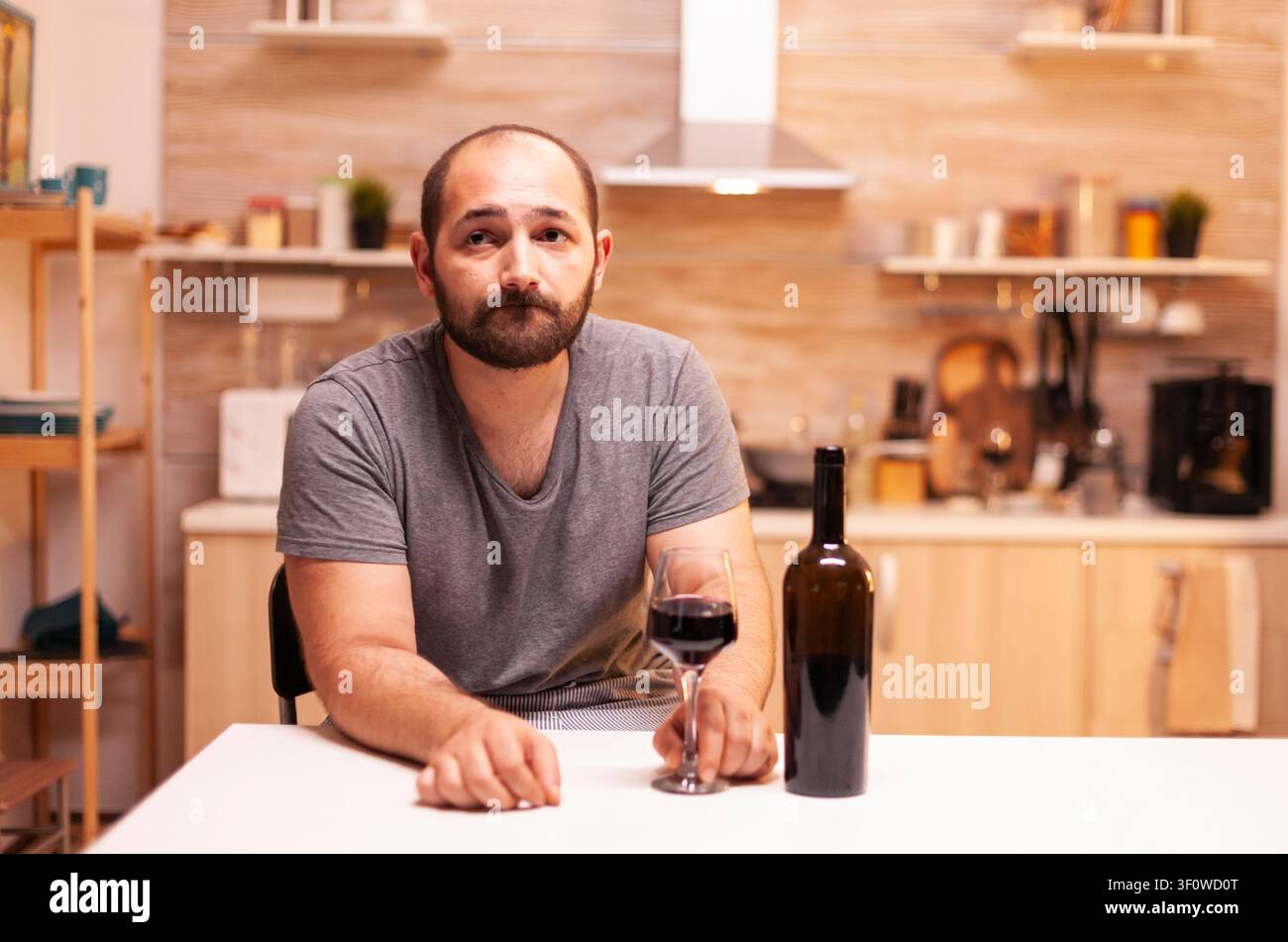 Giovane uomo pensieroso in un ambiente domestico che tiene un bicchiere di vino rosso e riflette sui problemi della vita. Persona infelice con sensazioni di malattia e ansia, esausta per problemi di alcolismo. Foto Stock