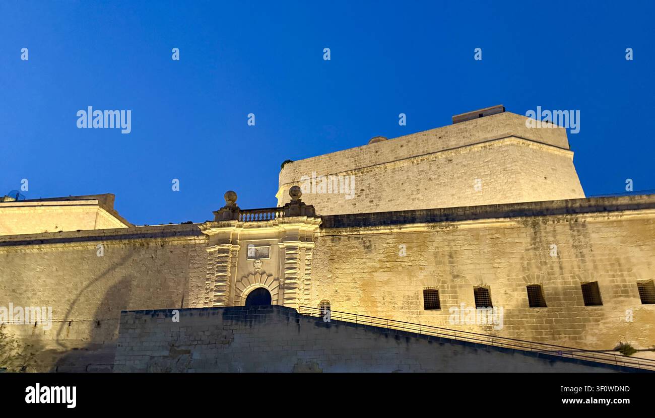 I riflettori dorati illuminano le mura della bastione in calcare e il portale di una fortificazione maltese contro un profondo cielo blu della sera. - Immagine stock catturata con smartphone