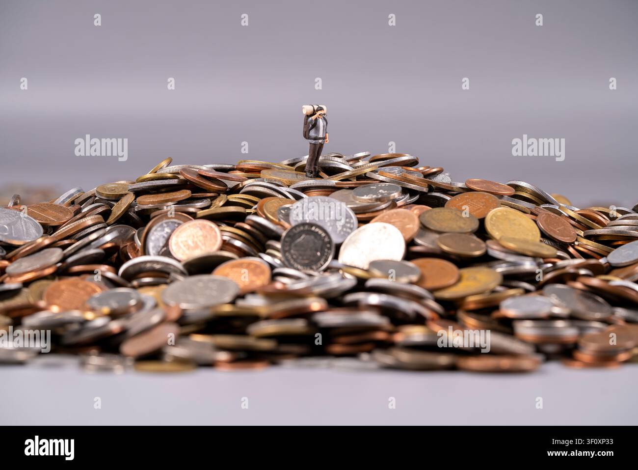 Una piccola figura si erge su una montagna di monete miste, mostrando una rappresentazione artistica unica di ricchezza e finanza. La scena enfatizza l'abbondanza Foto Stock