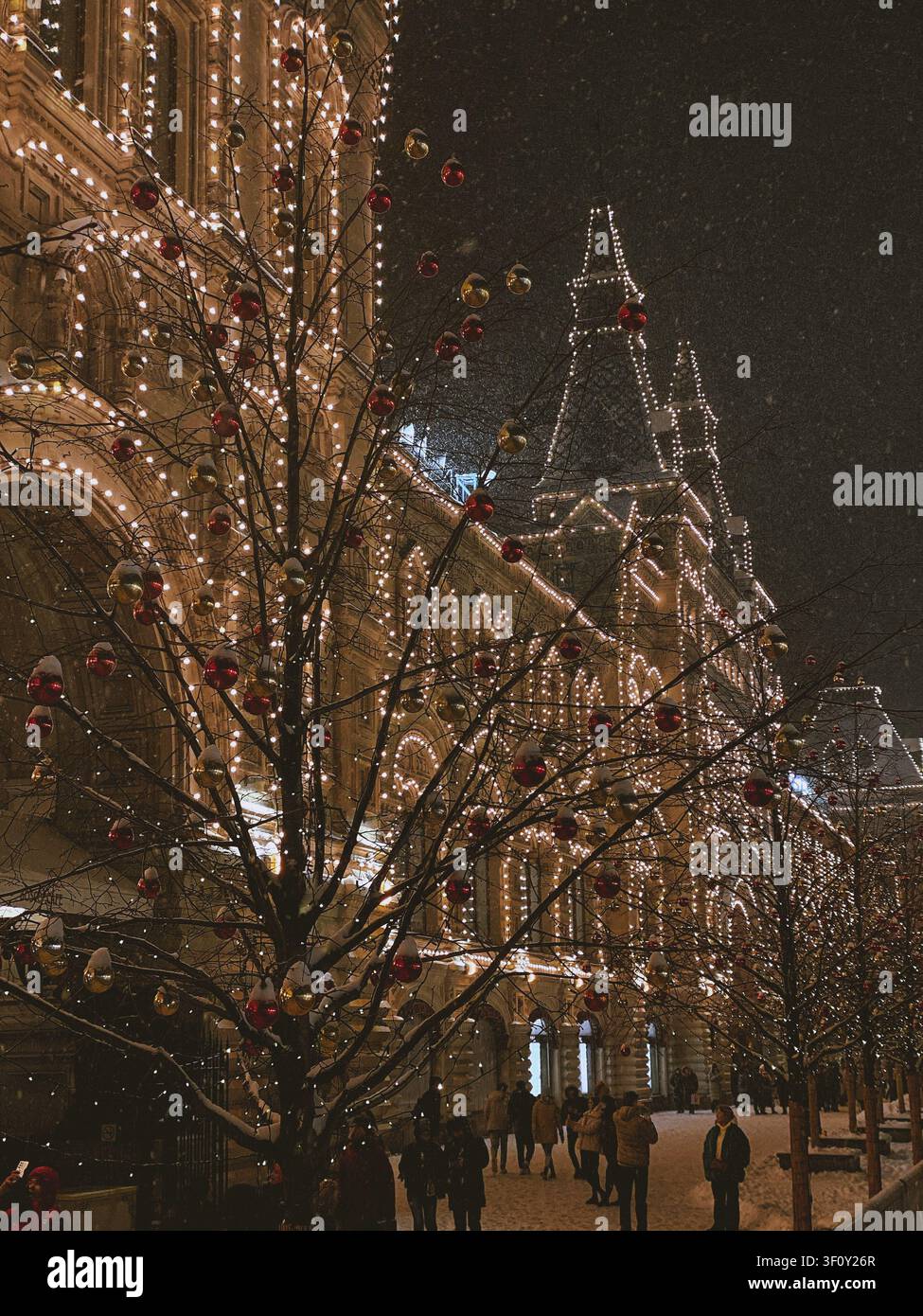 Mosca, Russia - 22 gennaio 2022: Alberi decorati a festa vicino a un edificio storico illuminato durante una notte invernale innevata - Immagine stock catturata con smartphone