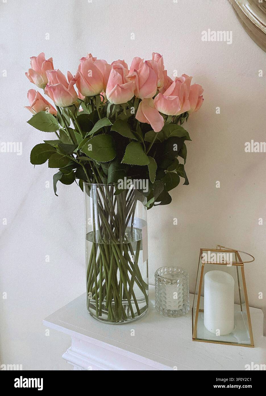 Rose rosa fresche disposte in un vaso di vetro trasparente accanto a candele decorative in un interno minimalista luminoso con luce naturale. Foto Stock