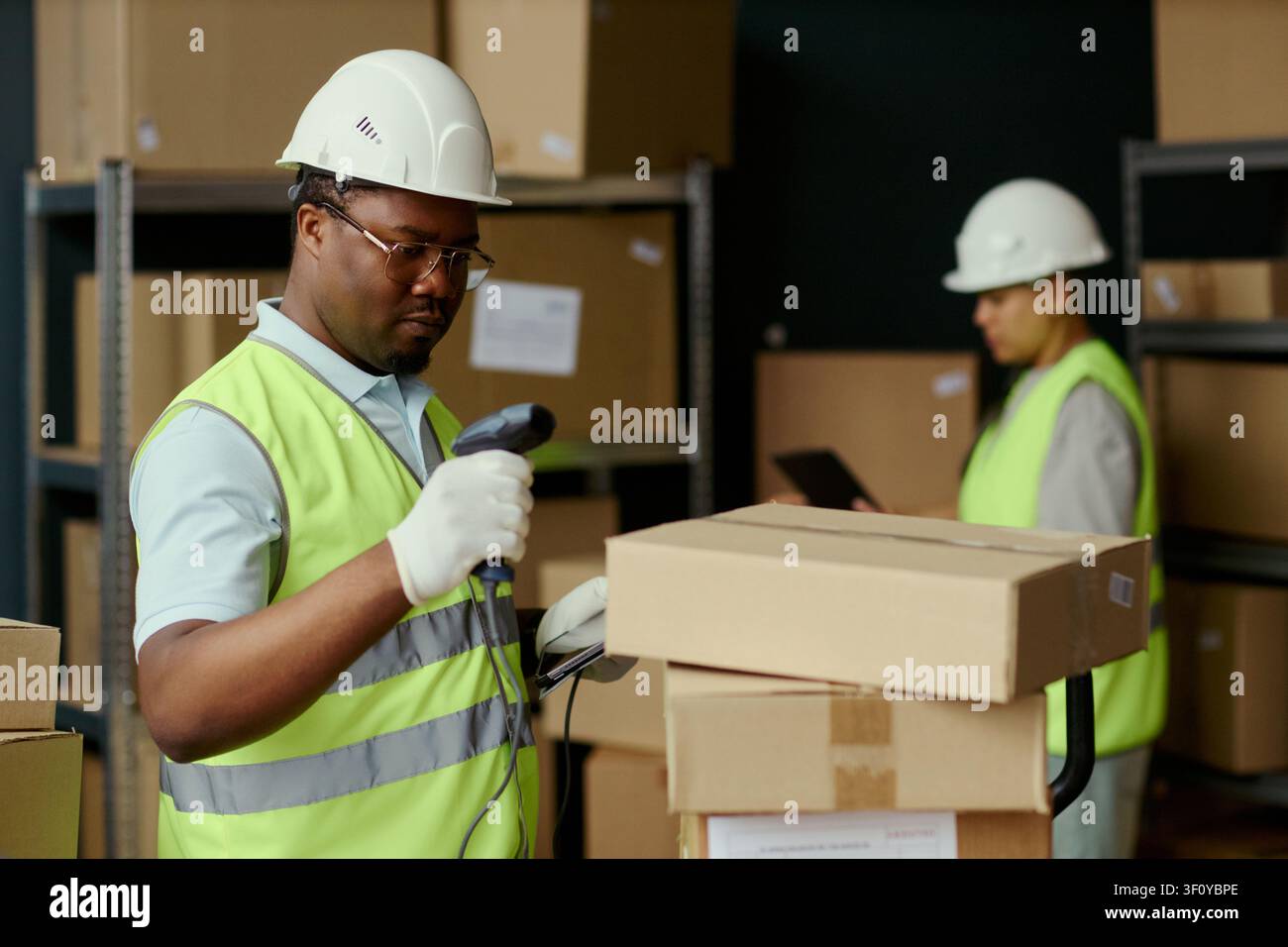 Scatto medio di un giovane addetto al magazzino di colore che scansiona il codice a barre di un pacco durante il controllo dell'inventario, con un collega che utilizza un tablet alle sue spalle. Ideale per campagne di logistica, gestione e spedizione Foto Stock