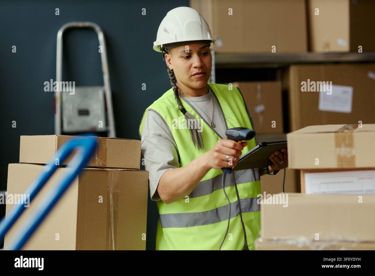 Prima media di una giovane donna biracial che scansiona pacchi con un lettore di codici a barre e controlla un tablet nel magazzino, gestendo l'inventario e l'adempienza. Ideale per logistica, catena di fornitura, e-commerce Foto Stock