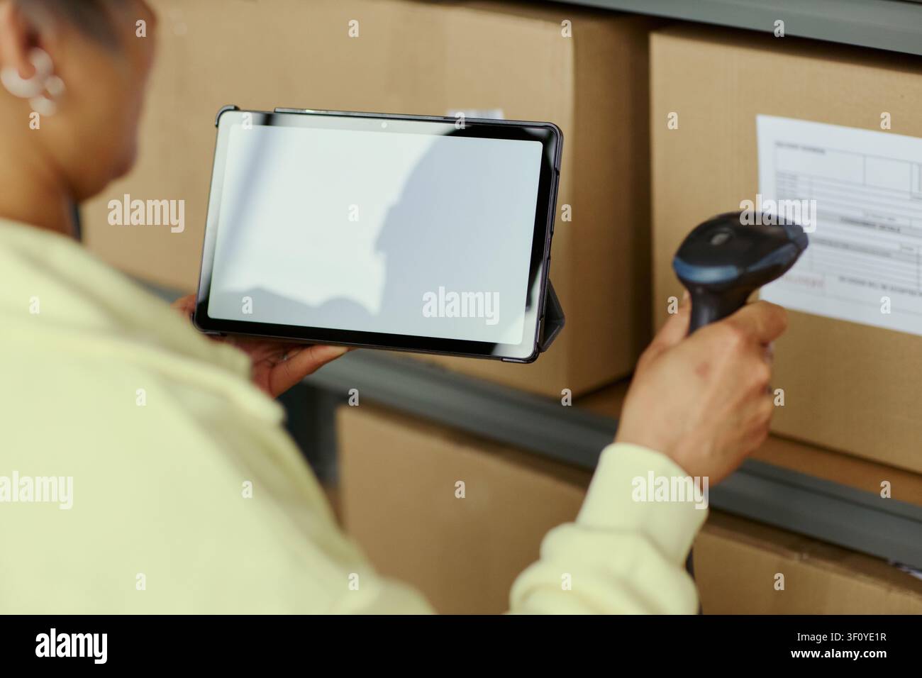 Close up di una giovane donna adulta che scansiona l'etichetta di una scatola con un lettore di codici a barre mentre controlla un tablet in un magazzino, gestendo l'inventario e il tracciamento degli ordini. Ideale per contenuti logistici e di e-commerce Foto Stock