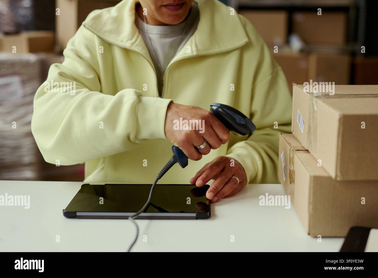 Primo piano di un giovane lavoratore adulto che scansiona il codice a barre di un pacco accanto a un tablet, elaborando una spedizione in magazzino. Ideale per l'adempimento e-commerce, la gestione dell'inventario, il marketing logistico Foto Stock