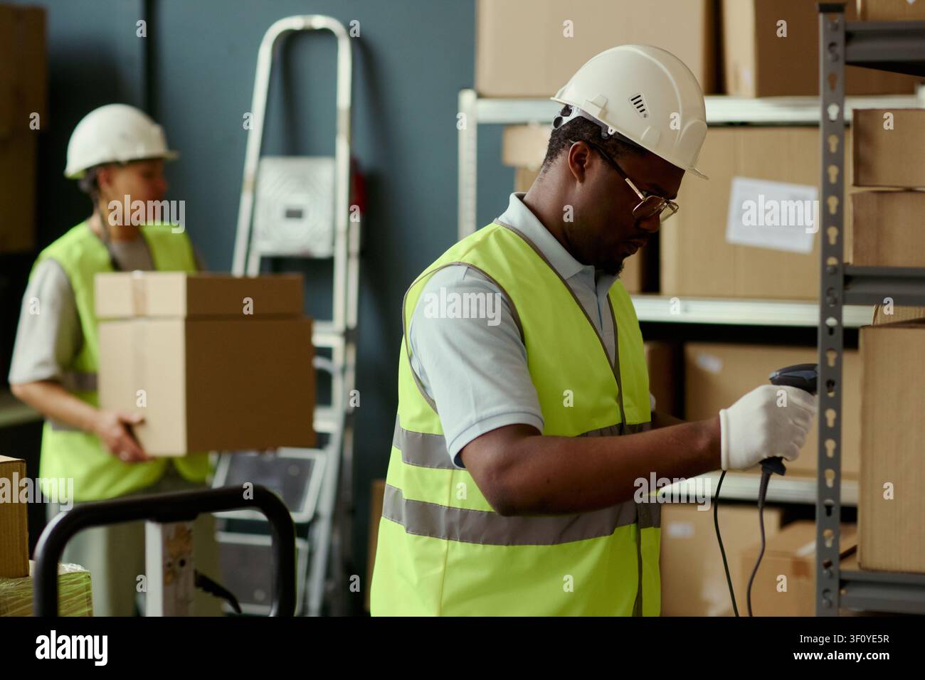 Primo piano di un addetto al magazzino di colore che scansiona l'inventario mentre una collega trasporta scatole nell'area di stoccaggio. Ideale per il personale logistico, la catena di fornitura, il marketing di adempimento Foto Stock