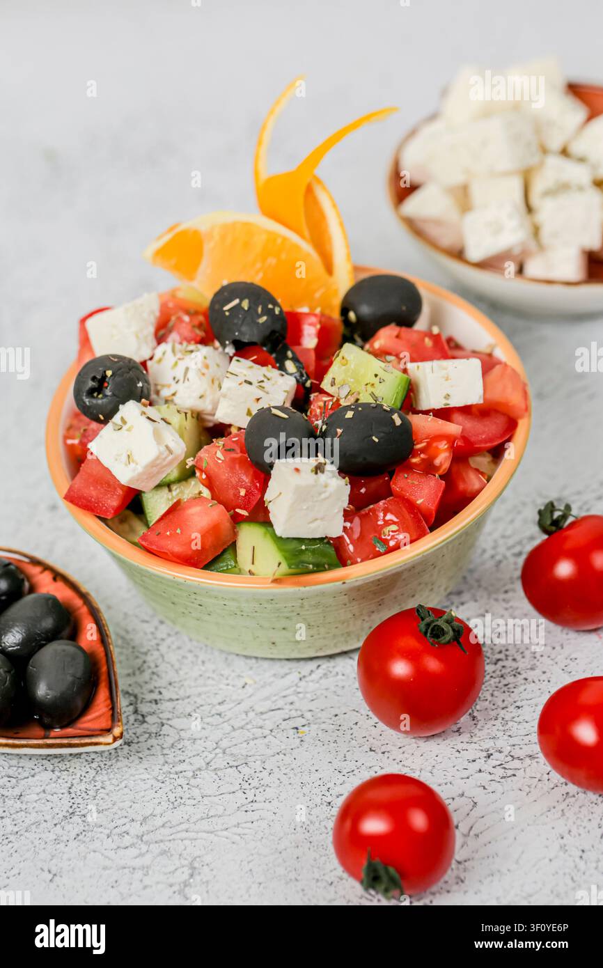 Una ciotola di insalata contiene pomodori a cubetti, cetrioli, olive e formaggio. Piccoli pomodori riposano nelle vicinanze. Una fetta di arancia aggiunge colore al piatto. Foto Stock