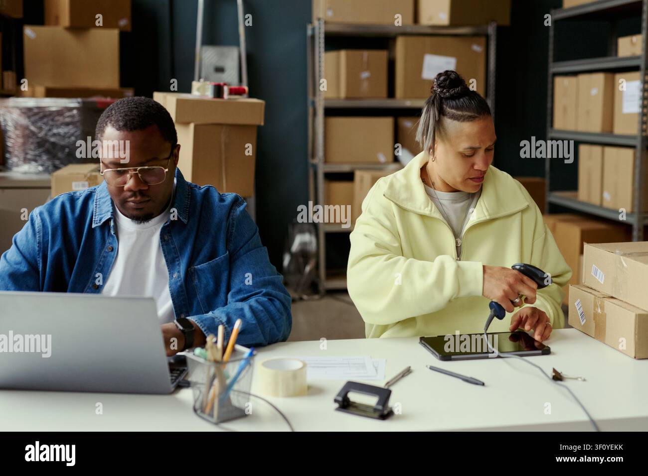 Scatto medio di un giovane afroamericano che digita su un laptop mentre una donna scannerizza etichette dei pacchi in un ufficio di magazzino, supportando il compimento e-commerce, il flusso di inventario, il marketing delle piccole imprese Foto Stock